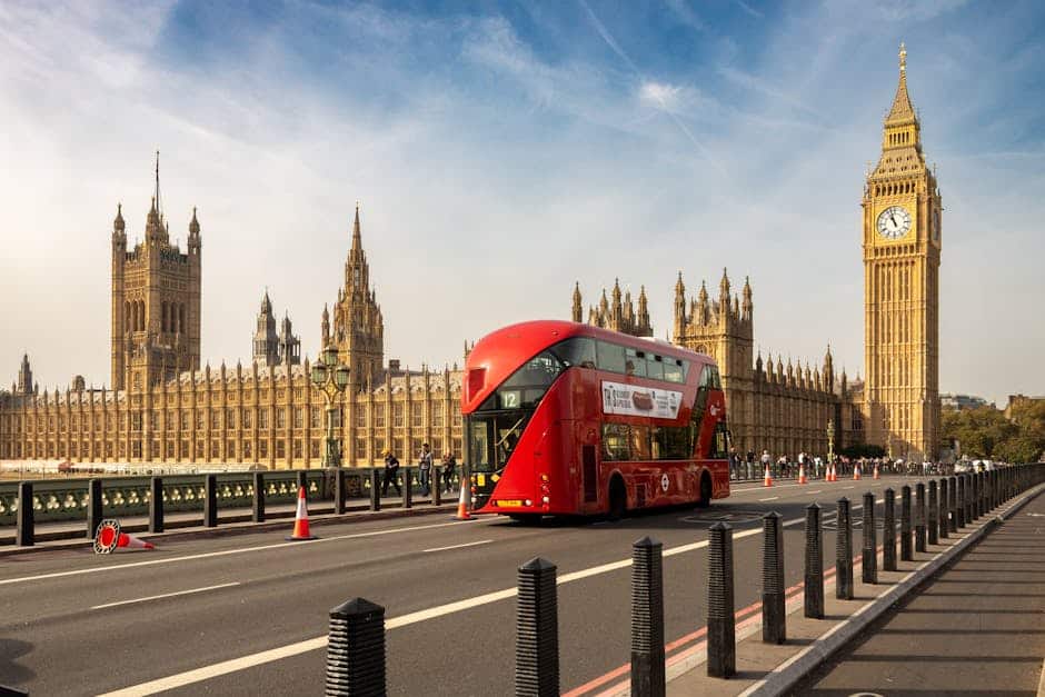 Sarkans Londonas divstāvu autobuss, kas brauc garām Big Ben un Parlamenta namiem zilu debesīm